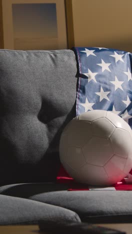 Vertikales-Video-Eines-Sofas-In-Der-Lounge-Mit-Amerikanischer-Flagge-Und-Ball,-Während-Sich-Die-Fans-Darauf-Vorbereiten,-Fußballspiele-Im-Fernsehen-Zu-Sehen